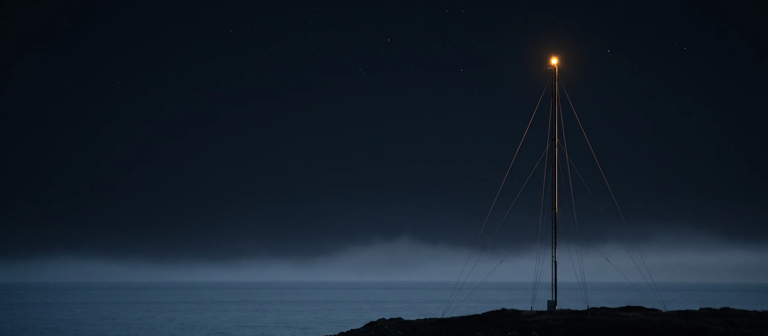A lone communications mast on a dark coastal headland at night, a single warm beacon lit at its top